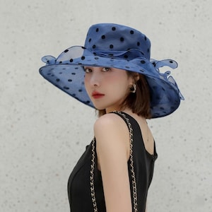 May include: A blue wide-brimmed hat made of a sheer, translucent fabric featuring a pattern of black polka dots. The hat has a structured crown and a ruffled, layered brim that creates a voluminous, wavy silhouette around the head.