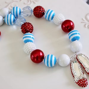 May include: A red, white, and blue beaded necklace with a pair of red jeweled shoes attached. The necklace features large, round beads with white and blue stripes, red beads, and clear beads.