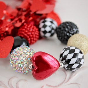 May include: A red heart-shaped bead, a clear rhinestone bead, a black and white checkered bead, a gold glitter bead, and a black bead. The beads are strung together on a necklace or bracelet.