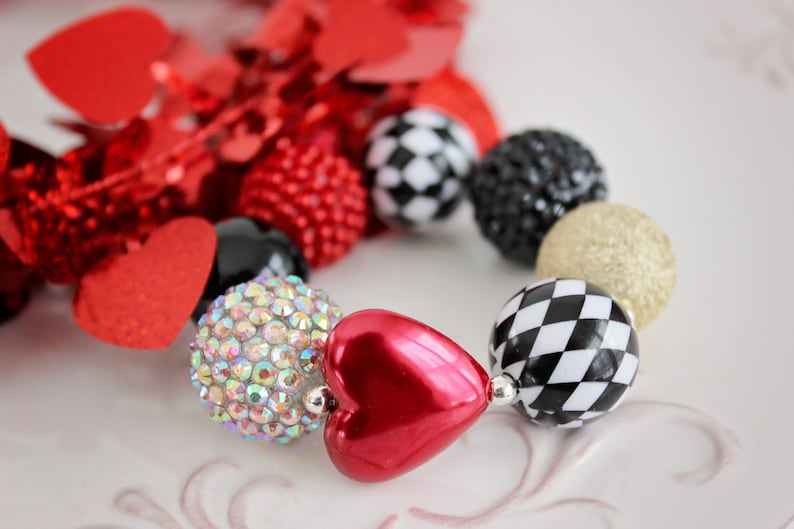 May include: A red heart-shaped bead, a clear rhinestone bead, a black and white checkered bead, a gold glitter bead, and a black bead. The beads are strung together on a necklace or bracelet.