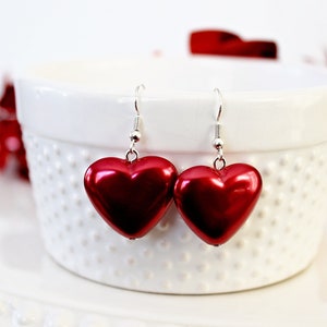 May include: A pair of red heart-shaped earrings with silver hooks. The earrings are hanging from a white ceramic bowl.