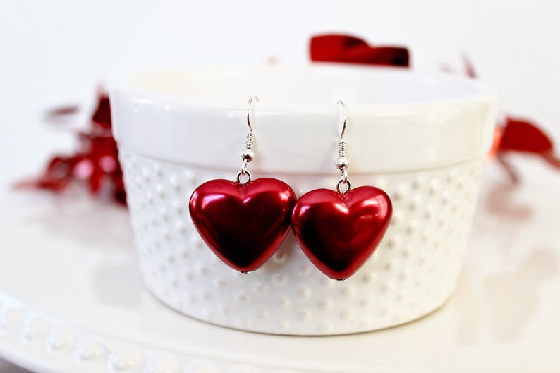 May include: A pair of red heart-shaped earrings with silver hooks. The earrings are hanging from a white ceramic bowl.