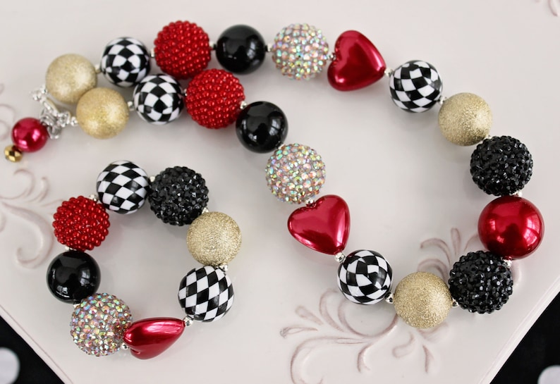 May include: A red, black, white, and gold checkerboard and heart-shaped bead necklace and bracelet set. The necklace and bracelet are made of large, chunky beads.