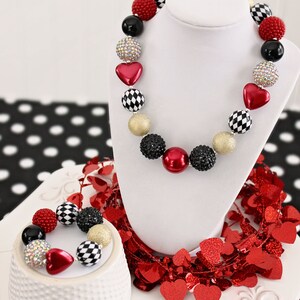 May include: A red, black, and gold beaded necklace and bracelet set with heart and checkerboard patterned beads. The necklace and bracelet are displayed on a white stand with red heart-shaped confetti.