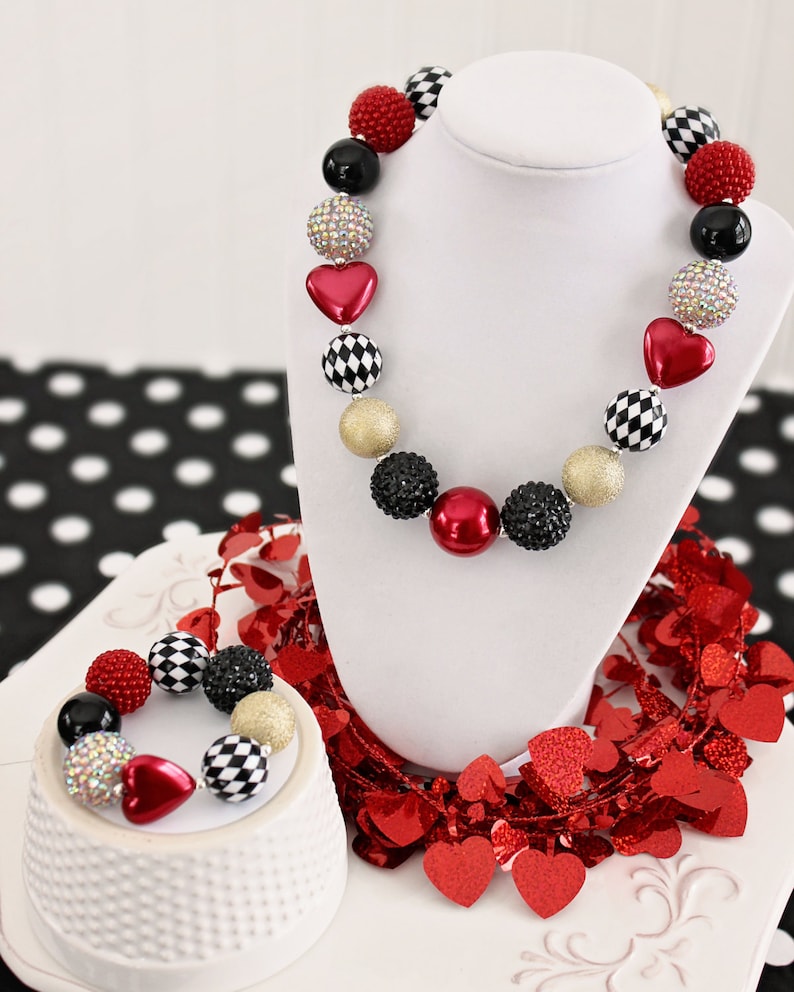 May include: A red, black, and gold beaded necklace and bracelet set with heart and checkerboard patterned beads. The necklace and bracelet are displayed on a white stand with red heart-shaped confetti.
