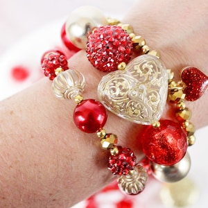 May include: A stack of bracelets with red and gold beads, including a large heart-shaped charm with a gold filigree design.