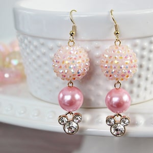 May include: A pair of dangle earrings with pink and iridescent beads. The bottom bead is a pink pearl and the top bead is a sparkly iridescent sphere. The earrings are finished with a gold-tone Mickey Mouse charm.