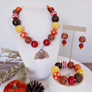 May include: A Thanksgiving-themed jewelry set with a necklace, bracelet, and earrings. The necklace features a turkey charm and is made with red, orange, yellow, brown, and white beads. The bracelet and earrings are made with similar beads.