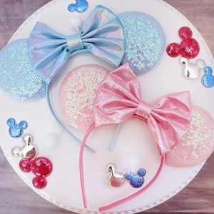 May include: Two pairs of iridescent Minnie Mouse ears with a headband. One pair is light blue with a light blue bow and the other pair is pink with a pink bow.
