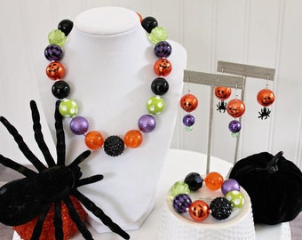 Puede incluir: Un conjunto de collar, pulsera y aretes con temática de Halloween. El collar y la pulsera presentan cuentas negras, naranjas, verdes y moradas con detalles de calabaza y araña. Los aretes son aretes colgantes con cuentas de calabaza naranja y dijes de araña negros.