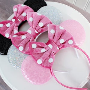 May include: Three pairs of Minnie Mouse ears with pink, silver, and black fabric. The ears are decorated with white polka dots and bows. The headband is pink.
