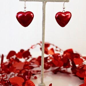 May include: A pair of red heart-shaped earrings hanging from a display stand. The earrings are made of a shiny, metallic material and have a simple, elegant design.