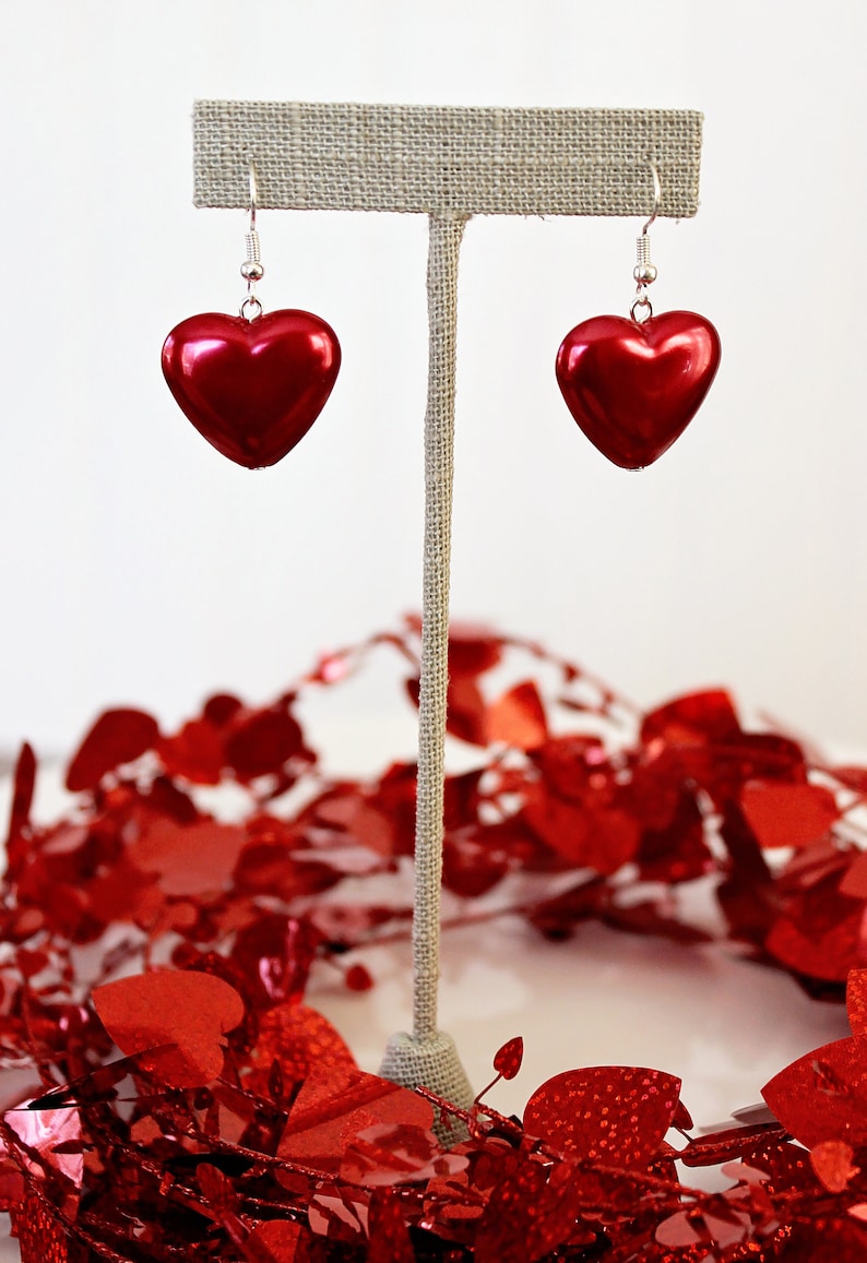 May include: A pair of red heart-shaped earrings hanging from a display stand. The earrings are made of a shiny, metallic material and have a simple, elegant design.
