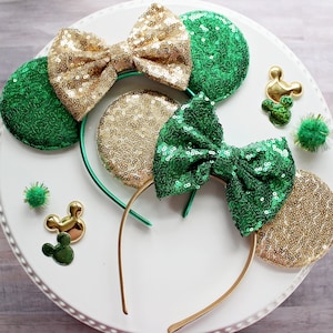 May include: A pair of gold and green sequined Minnie Mouse ears with a green bow. The ears are on a white background with gold and green Mickey Mouse heads.