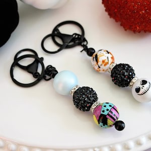 May include: Two black keychains with colorful beads. One keychain has a white bead with a black and white Jack Skellington face. The other keychain has a blue bead, a black bead with rhinestones, and a multi-colored bead with a patchwork design.
