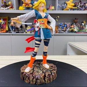 May include: A blonde anime character figurine wearing a blue and white kimono-style outfit with orange accents, holding a katana. The figure features a fox mask at the waist, black leg wraps, and orange leg warmers, standing on a textured brown rocky base.