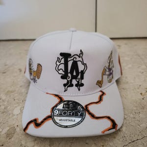 Könnte beinhalten: Eine weiße Baseballkappe mit einem schwarz gestickten LA-Logo und rauchartigen Umrissen. Die Kappe zeigt auf den Seiten gestickte Comic-Figuren sowie orangefarbene und schwarze Blitzmuster auf dem Schirm mit einem 9FORTY-Aufkleber.