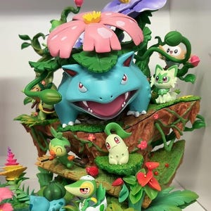 Puede incluir: Un diorama de varios niveles con un gran Venusaur azul con una flor rosa en la espalda, rodeado de figuras más pequeñas que incluyen a Rowlet, Sprigatito, Grookey, Chikorita, Snivy y Bulbasaur, todo sobre un entorno de follaje verde esculpido.