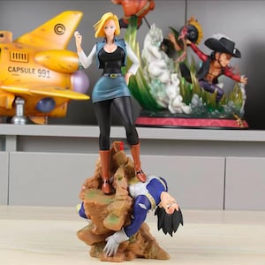 Puede incluir: Una figura de anime coleccionable con un personaje rubio con top negro, falda azul y botas marrones de pie sobre una base rocosa encima de un personaje derrotado con traje azul y blanco. Un avión amarillo Capsule 991 es visible al fondo.