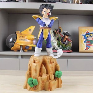May include: A figurine of a character with spiky black hair, wearing blue and white armor with yellow shoulder pads and a red scouter over one eye. The figure stands with arms crossed on a tan rocky base featuring small green trees.