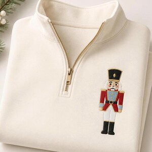 May include: A cream-colored quarter-zip pullover sweatshirt featuring a gold-toned metal zipper and an embroidered nutcracker figure on the chest. The nutcracker is depicted in a red, blue, white, and black uniform with a tall black hat.
