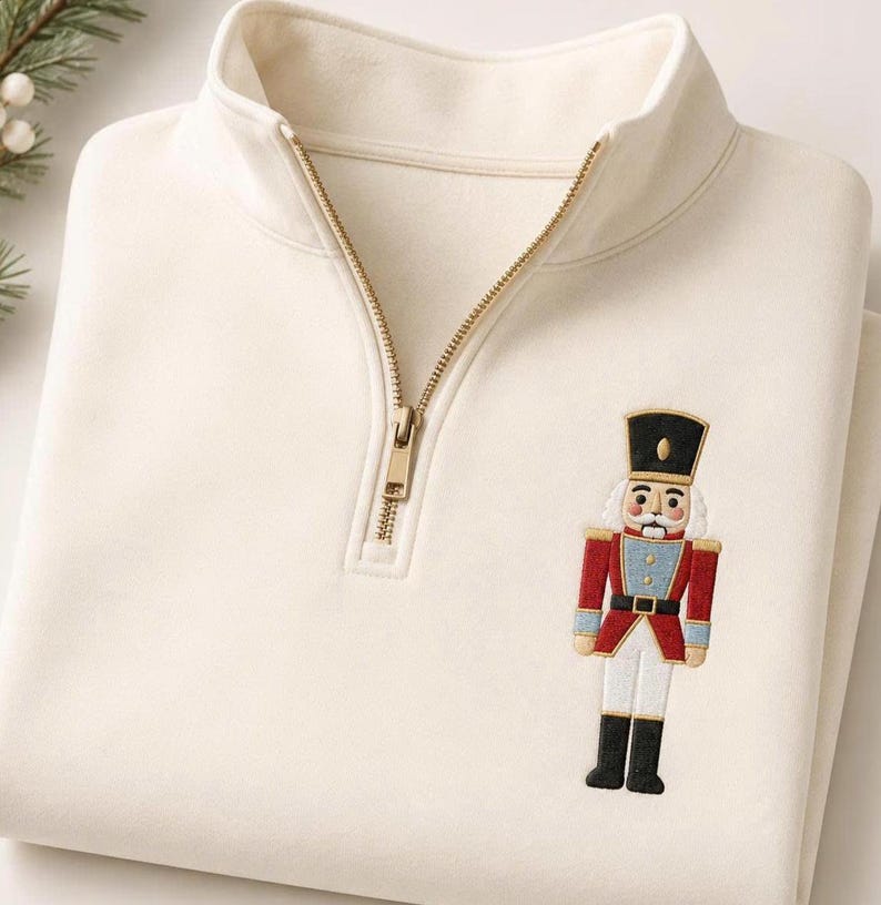 May include: A cream-colored quarter-zip pullover sweatshirt featuring a gold-toned metal zipper and an embroidered nutcracker figure on the chest. The nutcracker is depicted in a red, blue, white, and black uniform with a tall black hat.