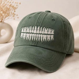 May include: A dark olive green baseball cap featuring an embroidered graphic of a full set of human teeth in white thread across the front panels. The cap has a curved brim, a button on the top, and stitched eyelets for ventilation.