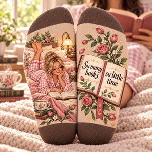 May include: A pair of beige socks with brown toe and heel sections. The left sock features a person in pink pajamas reading in bed with a lamp and tea. The right sock displays an open book with the text So many books so little time, surrounded by pink roses and a bookmark labeled Amelia.