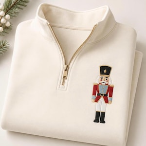 May include: A cream-colored quarter-zip pullover featuring a gold-toned metal zipper and an embroidered nutcracker figure on the chest. The nutcracker wears a black hat, a red jacket with blue accents, white trousers, and black boots.
