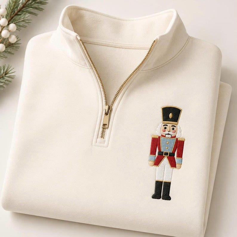 May include: A cream-colored quarter-zip pullover sweatshirt featuring an embroidered nutcracker figure on the chest. The nutcracker is depicted in a red, blue, white, and black uniform with a tall black hat and gold-toned zipper hardware.