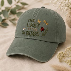 May include: An olive green baseball cap featuring embroidered text that reads THE LAST OF THE BUGS in dark green. The design includes a small yellow butterfly, a yellow dragonfly, a red ladybug, a white daisy, and green leafy sprigs.