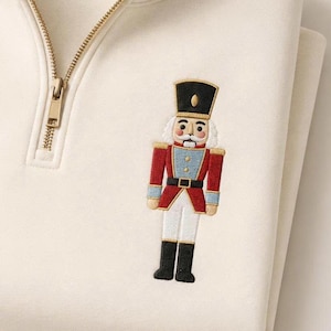 May include: A cream-colored quarter-zip sweatshirt featuring an embroidered nutcracker design on the left chest. The nutcracker wears a black hat, a red jacket with blue accents, a black belt, and white trousers with black boots. The zipper is gold.