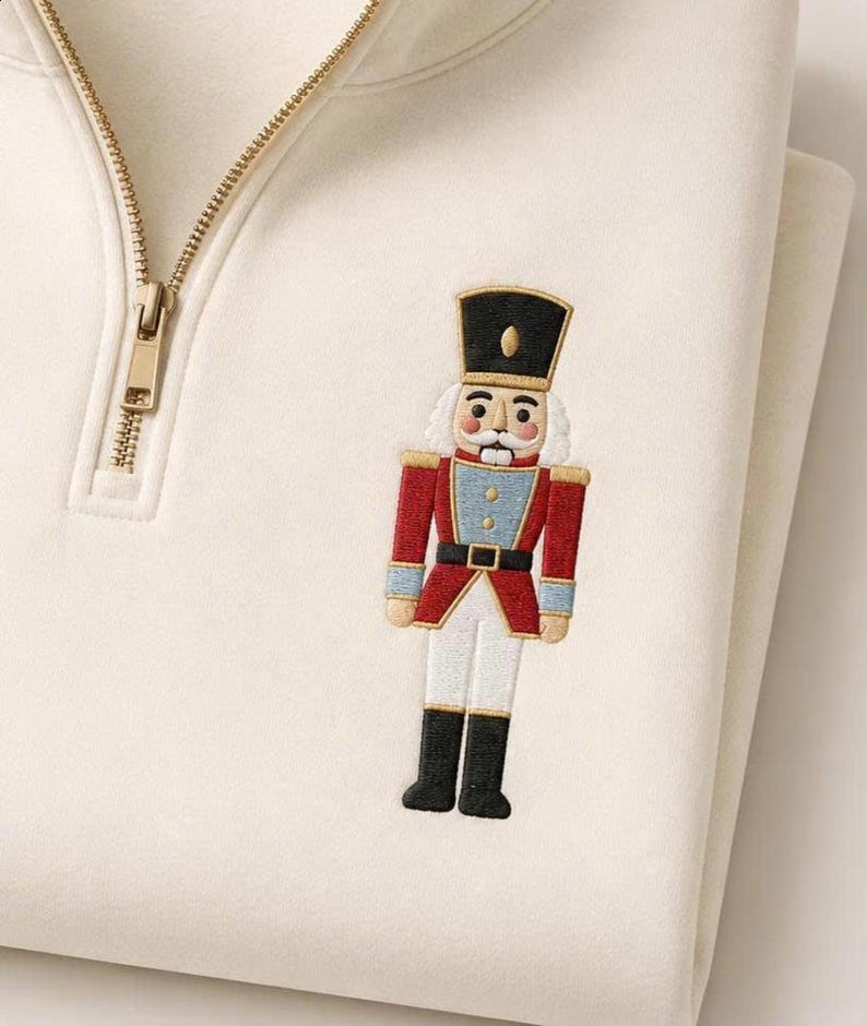 May include: A cream-colored quarter-zip sweatshirt featuring an embroidered nutcracker design on the left chest. The nutcracker wears a black hat, a red jacket with blue accents, a black belt, and white trousers with black boots. The zipper is gold.