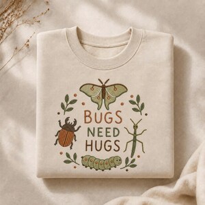 May include: A beige crewneck sweatshirt featuring an embroidered design of a moth, a beetle, a stick insect, and a caterpillar arranged in a circle. The text "BUGS NEED HUGS" is centered in brown letters, surrounded by small green leaves and dots.