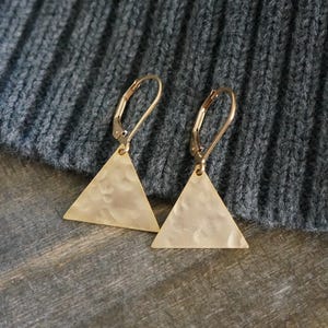 May include: A pair of gold-colored earrings with a triangular pendant. The earrings have a hammered texture and a lever-back closure. The background is a textured gray fabric and a wooden surface.