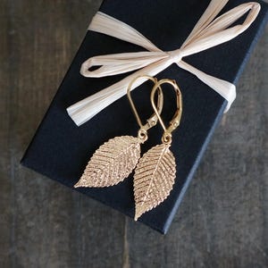 May include: Gold-colored leaf earrings with lever-back closures rest on a black gift box tied with a cream-colored ribbon. The earrings have detailed veining, and the box is on a dark, textured surface.