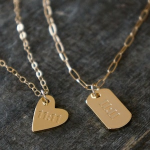 May include: Two gold-colored necklaces. One features a heart-shaped pendant with the engraved number 11:11. The other has a rectangular dog tag pendant with the same number. Both necklaces have delicate chains.