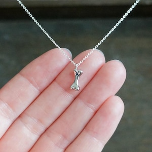 May include: A delicate silver-toned necklace featuring a small, polished dog bone-shaped pendant suspended from a thin, matching metal chain. The pendant has a smooth, reflective surface and is centered on the chain, creating a simple, clean look.