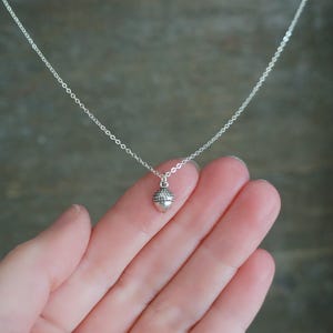 May include: A delicate silver necklace featuring a small acorn pendant. The acorn charm has a textured cap and a smooth body, suspended from a fine chain. The necklace is shown against a neutral background.