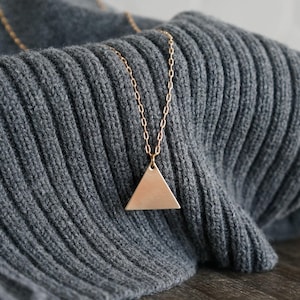 May include: A rose gold-toned necklace featuring a simple, flat triangular pendant suspended from a delicate, thin metal chain. The pendant has a smooth, matte finish and hangs centered against a textured, dark gray ribbed knit fabric.