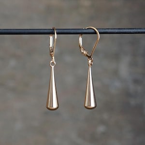 May include: A pair of gold-colored, teardrop-shaped earrings. Each earring has a lever-back closure and a smooth, polished finish. The earrings are suspended from a black rod against a blurred gray background.