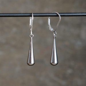 May include: A pair of silver-colored, teardrop-shaped earrings. Each earring has a lever-back closure and a smooth, polished finish. The earrings are suspended from a dark, horizontal bar against a blurred background.