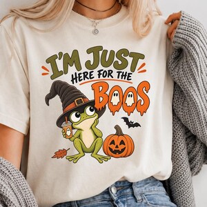 Puede incluir: Una camiseta de color crema con el gráfico de una rana verde con sombrero de bruja, sosteniendo una taza de café y sentada junto a una calabaza naranja. El texto dice I'M JUST HERE FOR THE BOOS en verde y naranja, con un pequeño murciélago negro.