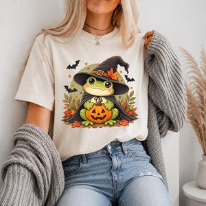 Op de afbeelding: Een crèmekleurig t-shirt met een afbeelding van een groene kikker die een zwarte heksenhoed en cape draagt en een kleine oranje pompoen vasthoudt. Het ontwerp bevat herfstbladeren, kleine sterren en zwarte silhouetten van vleermuizen.