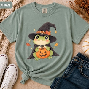 Puede incluir: Una camiseta de manga corta color verde espuma de mar con el gráfico de una rana verde claro que lleva un sombrero de bruja negro y una capa negra. La rana sostiene una calabaza naranja. Hojas de otoño y estrellas amarillas rodean a la rana.