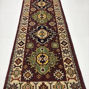 May include: A long, rectangular runner rug featuring a deep burgundy background with a series of repeating geometric medallions in shades of sage green, navy blue, yellow, and cream. The border displays intricate patterns with fringe at the ends.