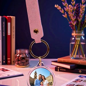 Peut inclure: Un porte-clés circulaire présentant une photo personnalisée d'une famille dans une place, attaché à une lanière en cuir texturé rose clair avec un bouton-pression en métal doré et un anneau fendu doré.