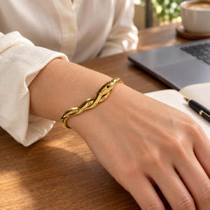 May include: A gold-toned snake-shaped bangle bracelet featuring a winding, textured body design. The bracelet is adorned with small, clear, round-cut stones embedded along the length of the snake, with a dark stone accent for the eye.