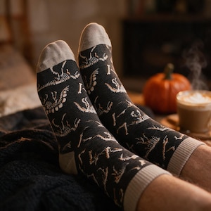 May include: A pair of black crew socks featuring a repeating pattern of white dinosaur skeletons. The socks have light grey heels, toes, and ribbed cuffs. The design displays various prehistoric skeletal poses across the dark fabric surface.