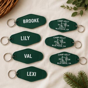May include: A selection of dark green keyrings with silver rings. Some keyrings display names such as "Brooke", "Lily", "Val", and "Lexi" in white text. Other keyrings say "Camp Kelly" with a tent graphic and "Blue Ridge, GA".